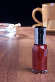 Red nail polish standing on a coffee table in front of a cup of coffee and an ashtray.