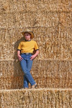Teen boy standing on straw staw looking into distance with yellow shirt and blue jeans with boots and hat on in the sun