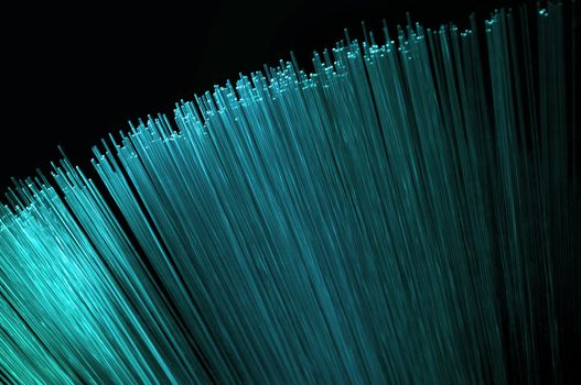 Abstract style close up on the ends of many bright cyan illuminated fiber optic light strands arranged over black.