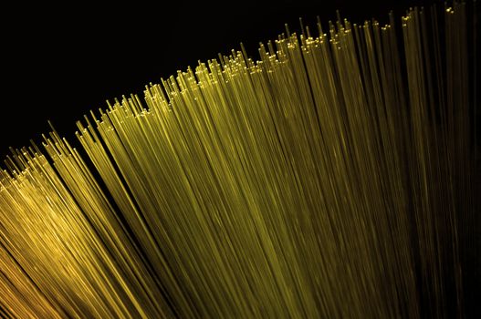 Close up on the ends of many illuminated golden fiber optic light strands arranged over black.