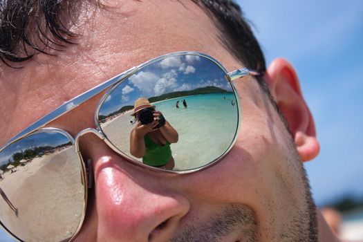 Close up of a man wearing reflective sunglasses in a tropical beach with reflection of the female photographer in the lens. Shallow depth of field.