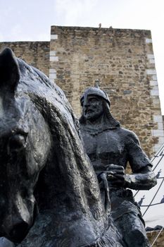 Statue of a medieval horseman warrior on top of a horse, behind a castle tower.