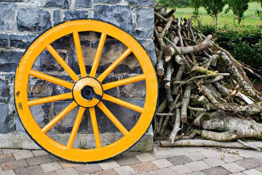 An old antique yellow wooden wagon wheel leaning against a stone wall. There is a pile of tree branches laying next to it.