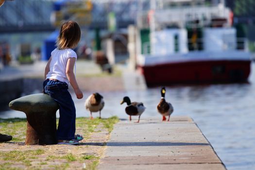 Little beauty girl feedind wild ducks at landing stage or river embankment at sunny day. She is wait for duck. We see ship not in fokus at background. Prague, Vltava.