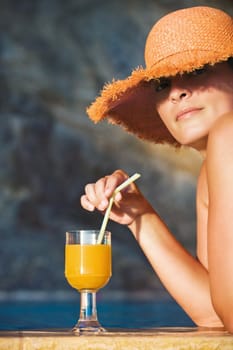 Attractive woman in hat ready to enjoy a glass of orange juice by the pool, looking at the camera
