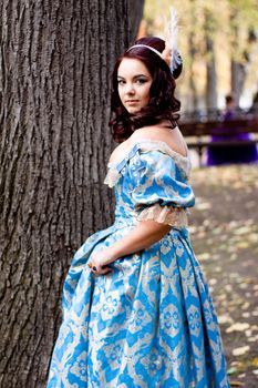 A portrait of lady in a blue baroque dress standing near a bole
