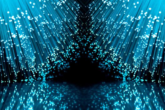 Two groups of blue illuminated fiber optic light strands against a black background and reflecting into the foreground.