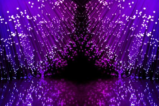 Close up on two groups of illuminated blue and magenta fiber optic light strands against a black background and reflecting into the foreground.