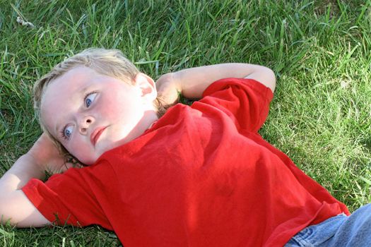 Little boy in red laying in the shady grass with hands behind head deep in thought
