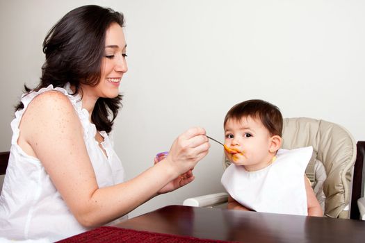 Beautiful happy mother or nanny feeds baby boy girl orange puree with spoon, infant eats messy, while sitting at table.
