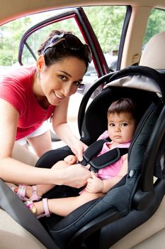 Happy smiling mother placing baby in car seat and closing belt for safety.