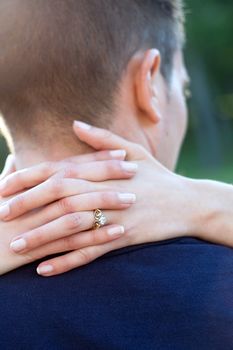 A happy woman hugs her fiance by placing her hands around his neck. Shallow depth of field with focus on the diamond engagement ring.