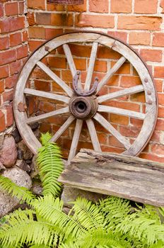 Antique wooden carriage wheel with binding of metal and horseshoe on it. Red brick wall. Fern leaves.