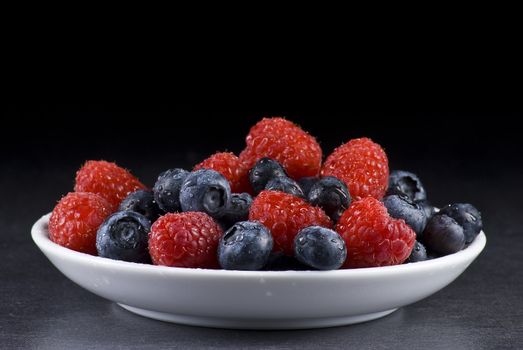 Plate of fresh blueberries and raspberries on balck background