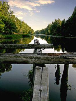 Ruins of wooden bridge on by a water smooth surface. Sunset of a sun.