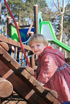 A blue-eyed small blonde toddler girl wearing a red coat is playing on a playground. Very colorful playground theme.