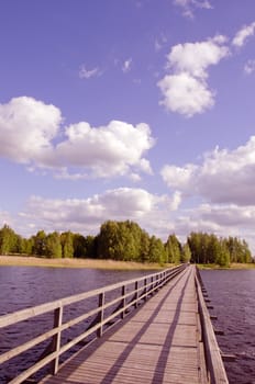 Long wooden footbridge with handrails over the lake and the man walking far away. Forest in the distance and the cloudy sky.