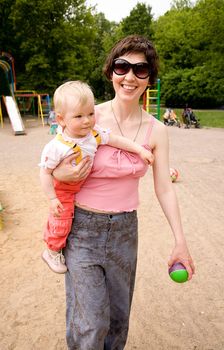 Beautiful harmonous mum with dark hair in sun glasses carries a little daughter and a green ball on a children's playground
