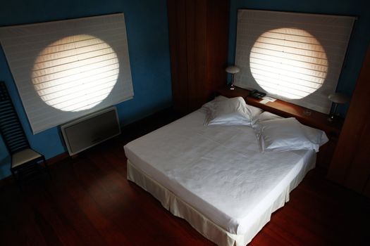 Bed with white blankets in bedroom with wooden floor and sunshades on round windows