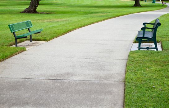 Two green metal benches across a large concrete pathway in a park