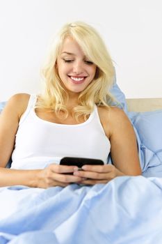 Smiling blonde woman texting on phone in bed at home