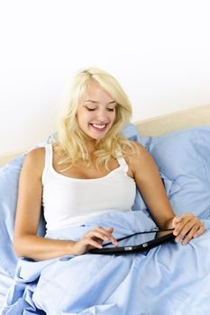 Happy blonde woman using tablet computer sitting in bed