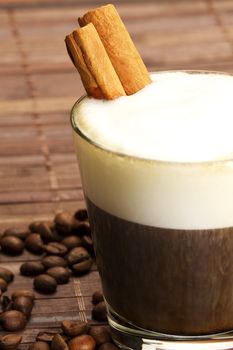 espresso coffee in a short glass with milk froth and cinnamon sticks inside and coffee beans on wooden background