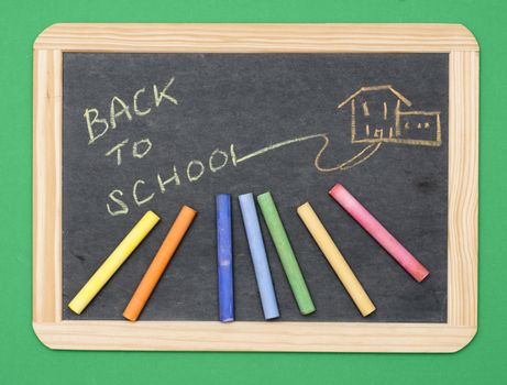 Overhead shot of small chalkboard with sketch of school and words Back to School written on it. Colored chalks also on surface. Green background enables image to be easily removed.