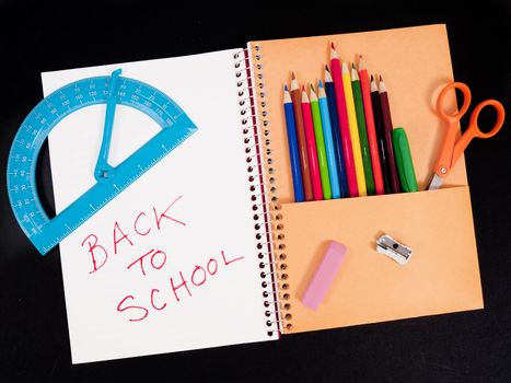 Back-to-school theme of student notebook with pocket full of colored pencils. Other items are scissors, protractor, sharpener, and eraser. Background is black.