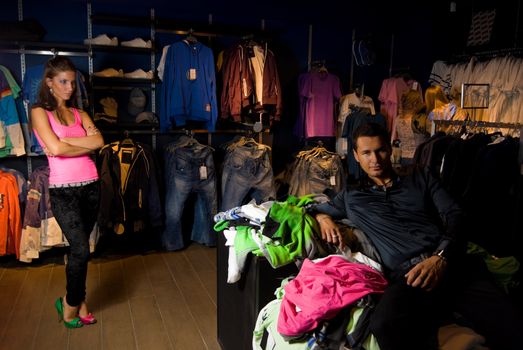 Young beauty is looking on her boy friend. He is laying on pile of shirts with blank face expression