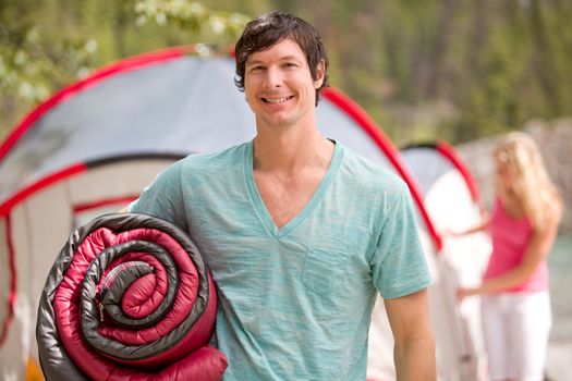 A portrait of a happy male camper holding a sleeping bag