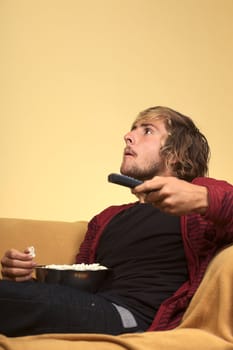 Young man watching TV holding a remote control in one hand and eating popcorn (Selective Focus, Focus on the left eye)