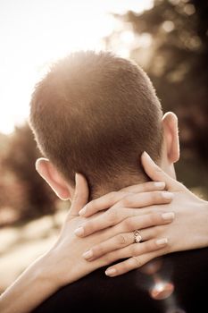 A woman hugs her fiance by placing her hands around his neck in sepia tone. Shallow depth of field with focus on the ring.