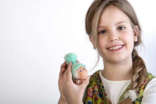 Studio portrait of a young blond girl who is showing her painted easter egg with a wool hat