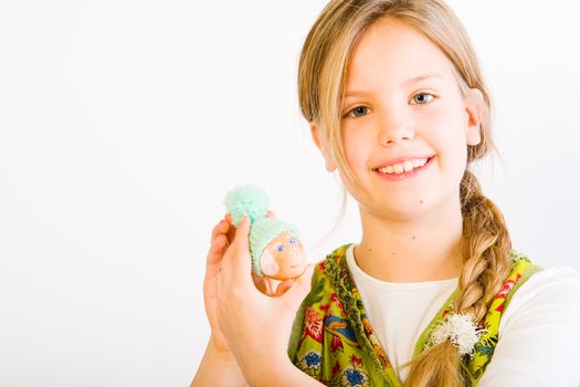 Studio portrait of a young blond girl who is showing her painted easter egg with a wool hat