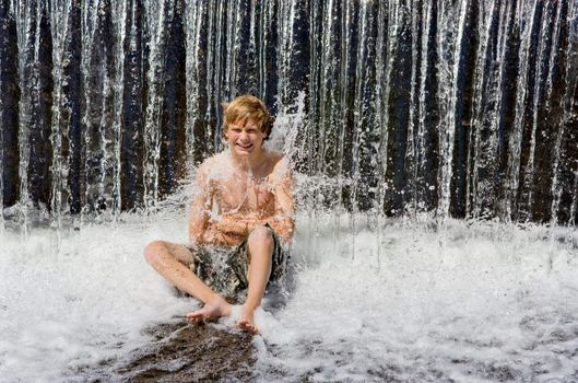 A boy sitting under a waterfall getting splashed by the water