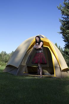 twenty something women wearing goth style clothes and holding a small baby doll, coming out of a tent with a great big smile
