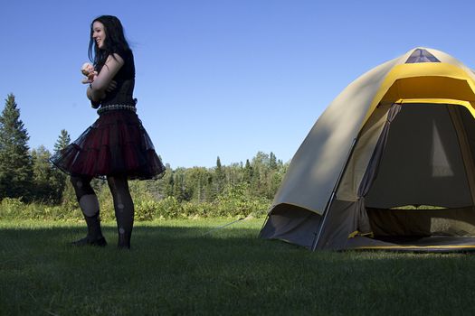 twenty something women wearing goth style clothes and holding a small baby doll, walking aways from a tent with a great big smile