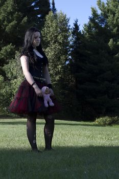 early twenty women wearing goth style clothes and holding a small baby doll, standing in a meadow with sad expression