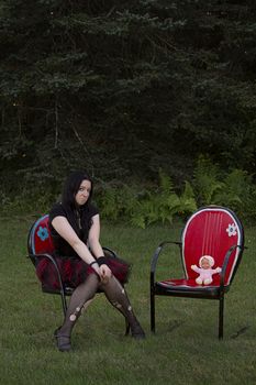 early twenty women wearing goth style clothes sitting on a lawn chair with her baby doll sitting in a lawn chair beside her