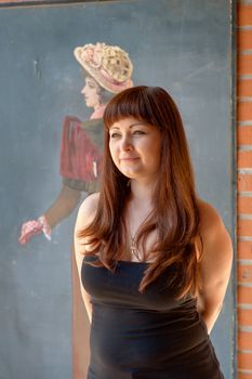 Young red-haired woman stands in front of old-fashioned drawing.
