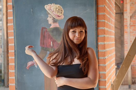 Young red-haired woman stands in front of old-fashioned drawing.
