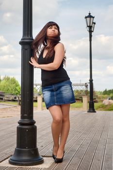 Young, sexy woman in short skirt leans on lantern pillar and looks upwards.
