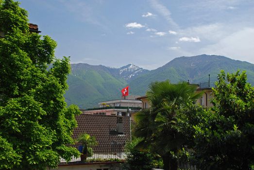 It is a point of view ftom the Lucarno ald city, there are building roofs, green trees, palms, Switzerland flag, mountains, blue sky