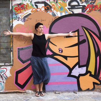A young girl stands before a wall covered with graffiti, arms outstretched