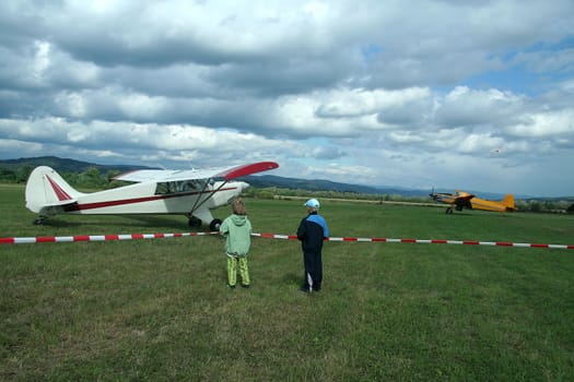 two kinds watching aeroplane preparing for start, another plane in background, and one just flying in distance