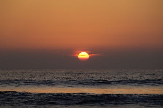 A picture of the red sun, as it sets over the calm ocean waters.