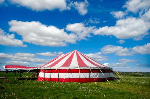 A red and white striped circus tent in green nature. The sky is blue with white cumulus clouds