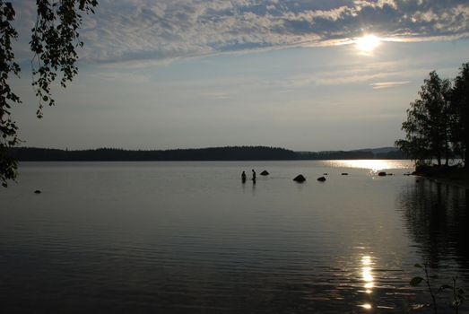 Waiting for a sunset at beutiful lake Kuuhankavesi in Central Finland, there are people in the water close to Ruokoniemi camping of Hankasalmi village, there are clouds and sun in the evening sky