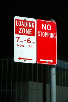 two red parking signs: LOADING ZONE and NO STOPPING, dark background, more text on signs